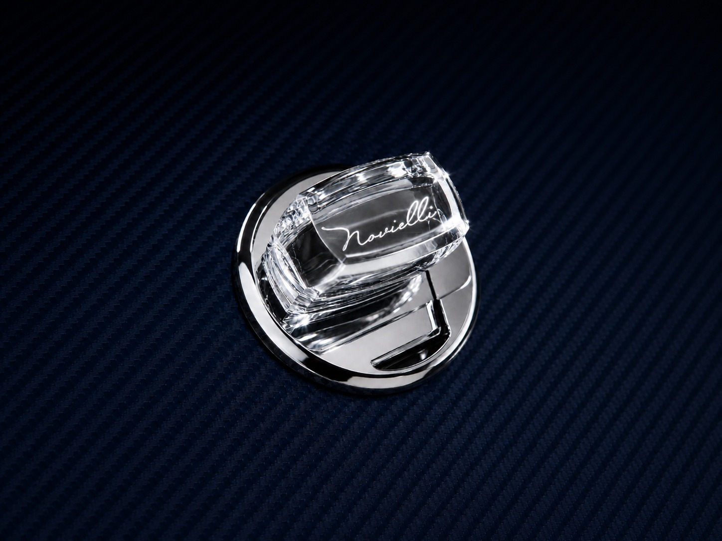 Polished chrome Novielli emblem with engraved script on a deep navy carbon-fiber surface.