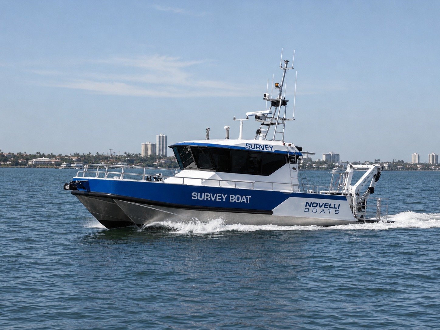 Novielli aluminum survey boat with blue hull underway on calm water with a coastal city skyline in the background.